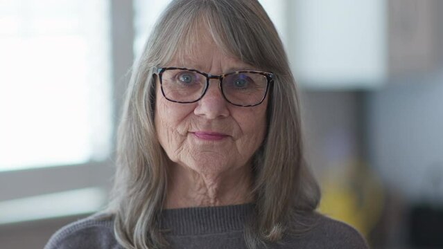Close up portrait of grey haired senior woman with glasses looking at camera. Older Caucasian retiree in a gray sweater. Slow motion 4k