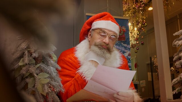 Santa Claus Reading Christmas Letter From Child. Saint Nicholas Getting Surprised And Laughing While Checking Christmas Mail At The Table. Merry Christmas Concept.