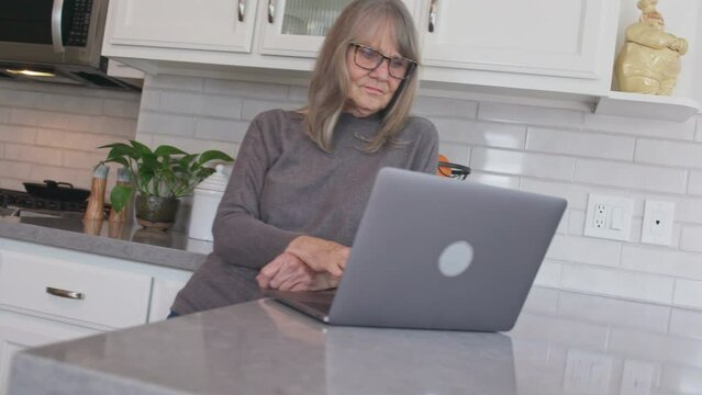 Low angle view of Senior white woman reading email on laptop computer in kitchen. Medium shot of older Caucasian lady working on pc at the counter. Slow motion 4k