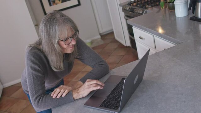 High angle view of Senior white woman working online from home on laptop computer in kitchen. Medium shot of older Caucasian lady working on pc at the counter. Slow motion 4k