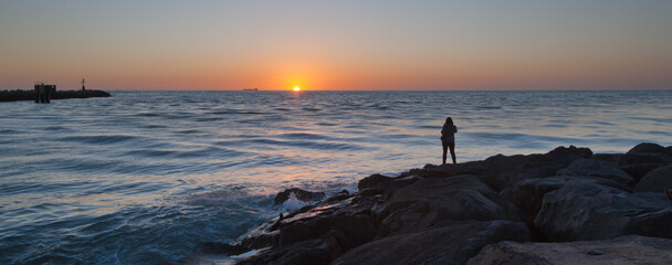 Sun setting on sea horizon with waves and foreground rocks and a silhouetted human figure watching the scene