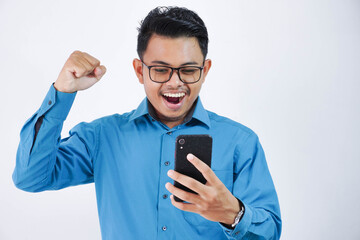 very excited young asian employee man with glasses holding phone with clenched fists wearing blue shirt isolated on white background