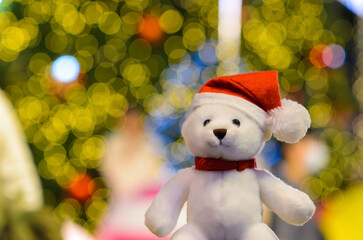 Santa claus teddy bear wearing hat sitting in front of colorful bokeh lights of Christmas tree.