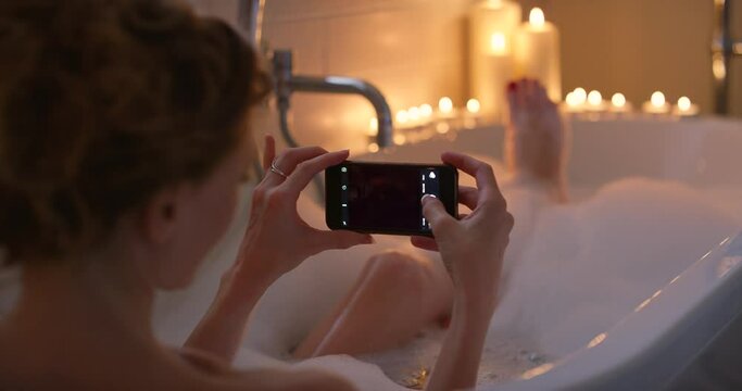 Woman, bath and phone picture, screen and relax, candles and cleaning body in bathtub, social media and calm. Female bathing, water and foam with mobile smartphone, hygiene while relaxing and peace