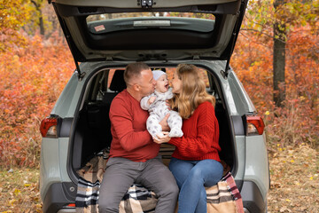 Parents in red sweaters sit in the trunk of a car and hug their baby