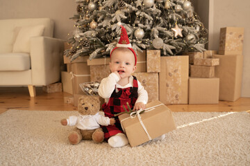 A cute baby in a Santa hat sits and gnaws his finger under a festive Christmas tree with a gift and a teddy bear