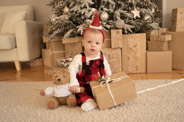 A cute baby in a Santa hat sits under a festive Christmas tree with a gift and a teddy bear