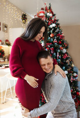 Husband listening to pregnant wifes stomach at Christmas in an apartment against the backdrop of a Christmas tree. Christmas morning. Woman in a red dress and a man in a gray sweater