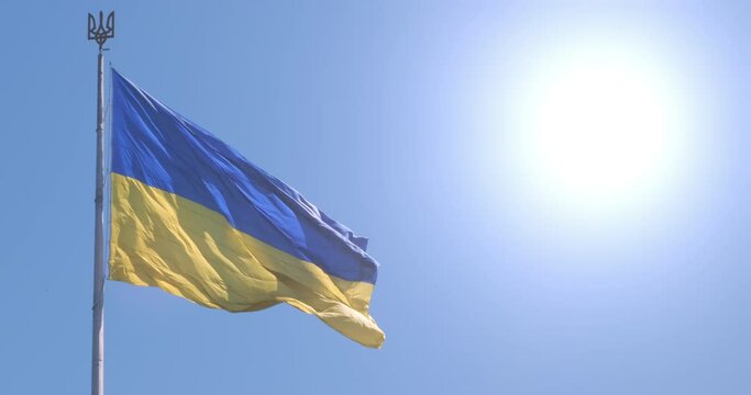 Yellow-blue Ukraine flag waving in sun light. Ukraine flag on sunny blue sky slow motion. Ukrainian flag wind swaying in sunlight. Tryzub coat on flagpole arms. National symbol background. 4k footage