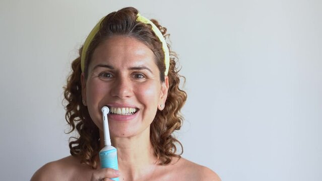 Middle-aged caucasian curly woman brushing teeth with electric toothbrush over white background. Morning routine personal hygiene. Blank for advertisement, mock-up, template