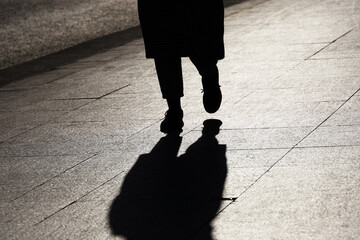 Black silhouette and shadow of lonely woman walking on a street. Female legs in boots on paved city sidewalk