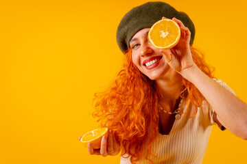 Redhead woman holding an orange over isolated yellow background, winking with the fruit