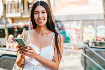 Optimistic Hispanic female in white dress with backpack browsing smartphone and looking at camera with smile on blurred background of sunlit street in Barcelona, Spain