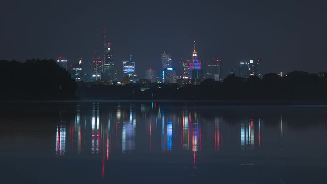 Night city skyscrapers skyline time-lapse. Warsaw and Vistula river. The lights of the night city are reflected in the water. Lightning sometimes appears in the sky and planes fly by.