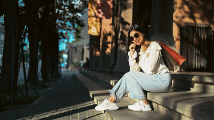 Beautiful hipster woman in trendy clothes and sunglasses sitting on street stairs after shopping