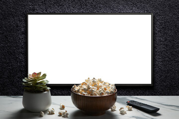 Television on a dark gray wall with remote control, home plant and popcorn bowl on the table. TV 4K...
