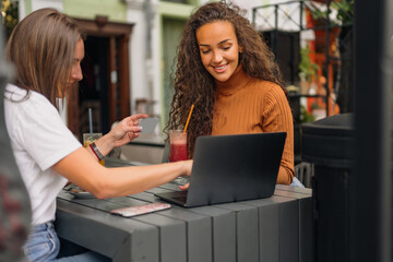 Two girls sitting in the cafe, one of the girls is holding a credit card while the other is typing something on the laptop.