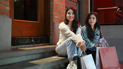 Photo of two pretty asian women in trendy clothes holding shopping bags and sitting on stairs at the shopping district of a city