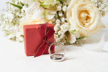 Two rings, different sizes, red box for rings, bridal bouquet on a white wooden background