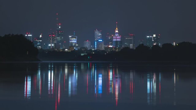 Night city skyline time-lapse. Night Warsaw / Warszawa skyscrapers cityline / downtown time lapse, over Vistula river. The lights of the night city are reflected in the water.