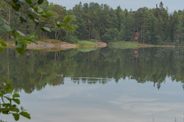 View of a quiet lake with a small red house on the shore surrounded by trees. Scandinavia. Natural background.