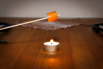 A piece of a sausage is grilled on a wooden skewer over the flame of a tea light placed on a beech...