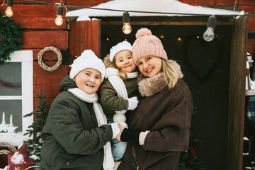 family mother, siblings girl sister and teenage cute boy brother in knitted sweater and hat having fun with first snow at porch of country house, concept of winter spirit and Christmas.