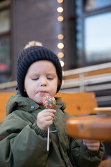 A boy in a green jumpsuit and a black hat sits on the veranda of a cafe on the street and eats sweets, cake, cake pop