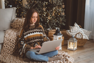 Portrait of smiling woman working on laptop or shopping online, buying new year presents with decorated Christmas tree on background, typing on keyboard.