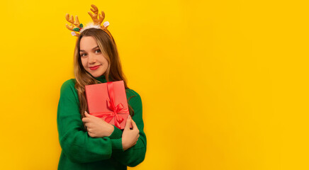 Happy smiling young woman holding pink present gift box with bow, wearing knitted green sweater cardigan with reindeer antlers headwear and looking at camera on yellow background with copy space.