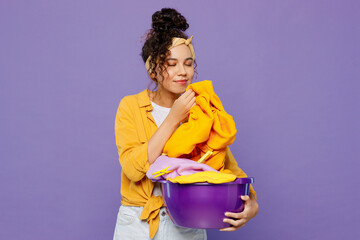 Young housekeeper woman wear yellow shirt tidy up hold basin with clean clothes after laundry sniff...