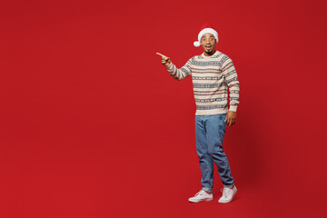 Fototapeta na wymiar Full body merry young man wear Christmas sweater Santa hat posing walk go point index finger aside on workspace area isolated on plain red background. Happy New Year 2023 celebration holiday concept.