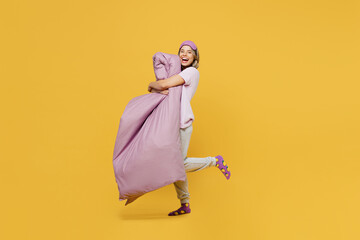 Full body side view happy young fun woman wears purple pyjamas jam sleep eye mask rest relax at home hold in hands blanket duvet isolated on plain yellow background studio portrait. Night nap concept.