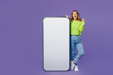 Full body fun young woman 30s she wear casual green knitted sweater stand near big huge blank screen mobile cell phone with area do winner gesture isolated on plain pastel purple background studio.