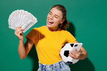 Young happy woman fan wear basic yellow t-shirt cheer up support football sport team hold in hand money soccer ball watch tv live stream use mobile cell phone isolated on dark green background studio. © ViDi Studio