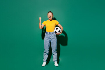 Naklejka na ściany i meble Full body fun young woman fan wears yellow t-shirt cheer up support football sport team hold soccer ball watch tv live stream do winner gesture celebrate clench fist isolated on dark green background.