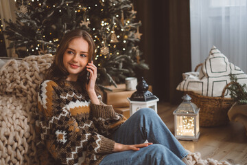 A charming young red-haired woman on the background of a Christmas tree in a cozy living room holds a mobile phone and waves her hand talks with her friend or family via video call