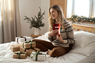 Pleased caucasian woman sitting on bed among Christmas present and hugging greeting card