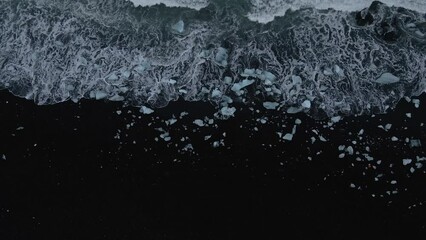 Waves with white foam breaking on black sand at Diamond beach in Iceland. Aerial ascending tilt-up reveal