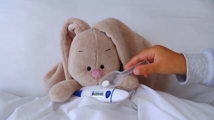 Child's little hand gives medicine a pill to a sick bunny hare toy lying in bed with a thermometer...