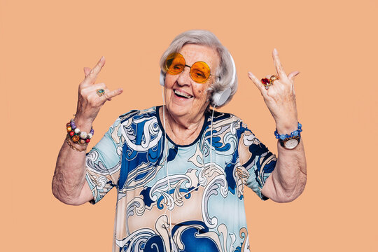 Elderly cheerful housekeeper grandmother woman 90s does horns sign gesture with hands. Stylish woman with hat, sunglasses, headphones and t-shirt over beige background.