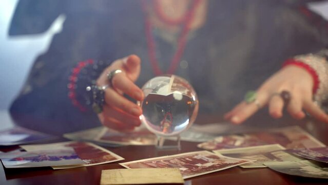 Female sorcerer gesturing above the round crystal ball and spelling in dark room with mist, scary witch reading potion, talking with spiritual ghosts and calling evil. Horror witchcraft, paranormal