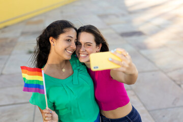 A beautiful lesbian young couple embraces and holds a rainbow flag. Girls taking selfie photo