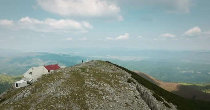 A drone shot of the pak of mountain Snežnik where is a mountain cottage and a hiker standind at by a rock monument. Mid range view around the mountain, sky with clouds.
