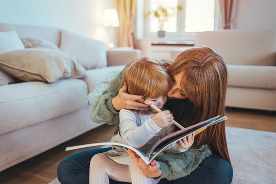 Caring smiling mother reading book with little kid girl lying on warm floor at home, mom or baby sitter playing having fun telling fairy tale to child daughter, underfloor heating, family activities