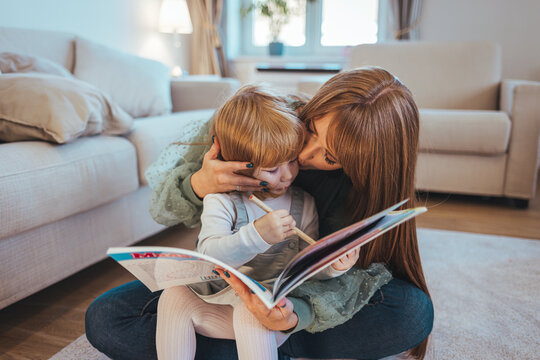 Single mother having fun at home with her baby daughter. Mother and daughter reading book on the floor. Mother reading a book to her daughter on carpet