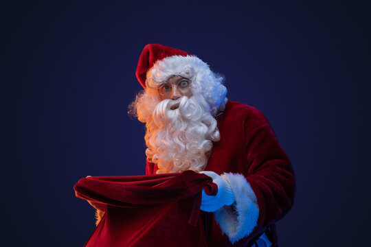 Portrait of bearded santa claus with red costume and bag against dark background.