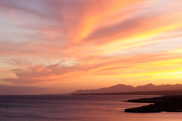 Fototapeta premium Horizontal seascape of sunset on the beach with silhouettes of mountains in the background and calm sea lapping the coast. Sky with orange, golden and yellow clouds. Lanzarote, Canary Islands, Spain. 