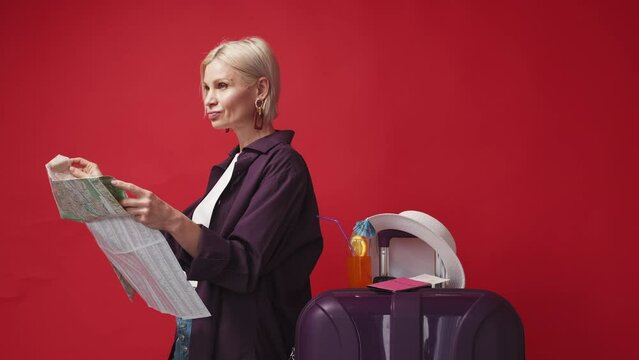 Choose destination. Inspired woman. Vacation journey. Smiling middle-aged lady choosing direction for travelling on map posing with suitcase on red background.