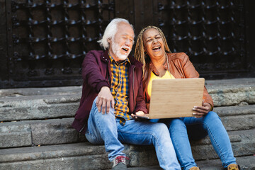Multiracial couple laughing with a video in the laptop sitting in a stairs outdoors. Happy mature...